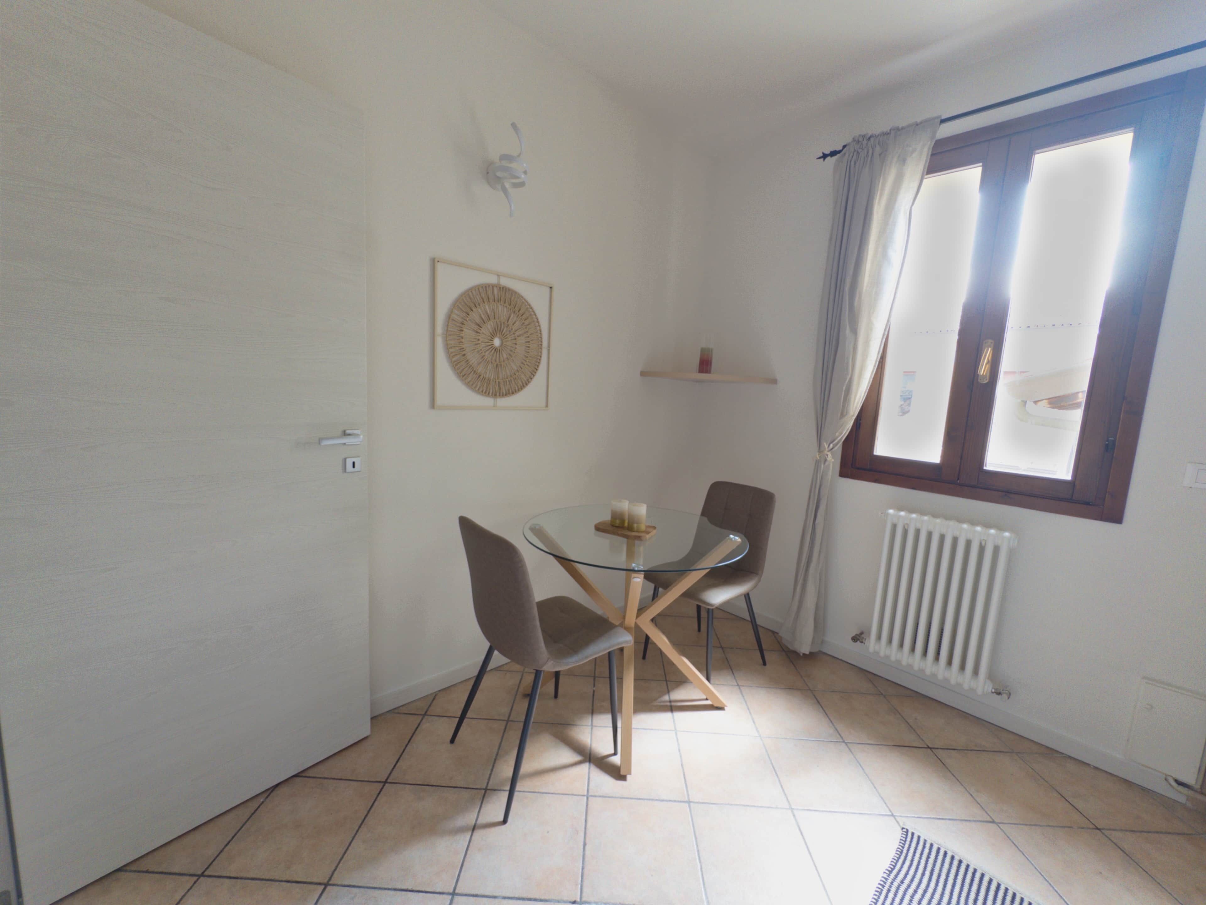 A room with a round glass table, four chairs, a wall-mounted light fixture, a framed artwork, a radiator, and a window with curtains.