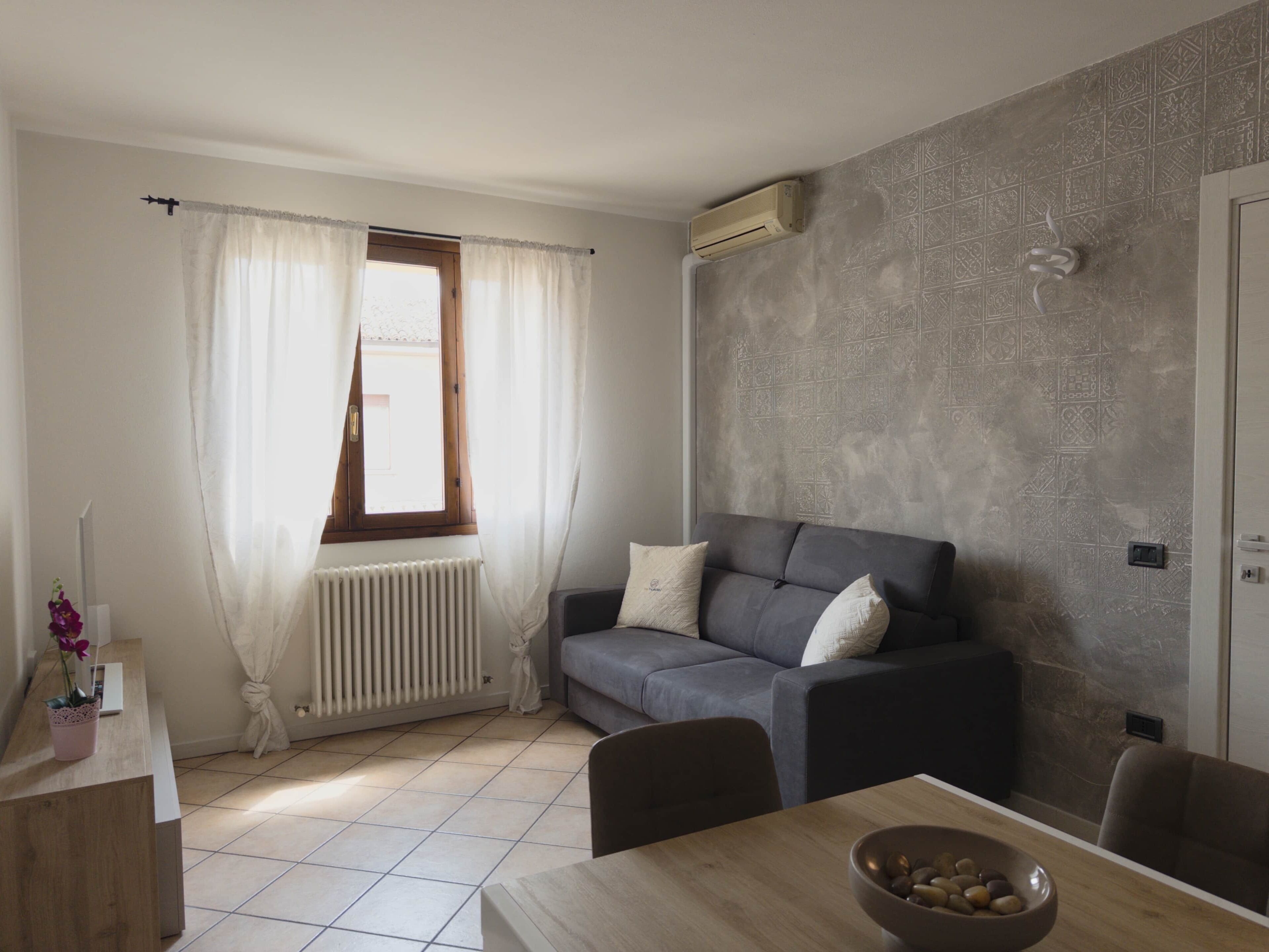 Een moderne woonkamer met een grijze bank, een eettafel met een schaal fruit, een radiator en een raam met gordijnen.