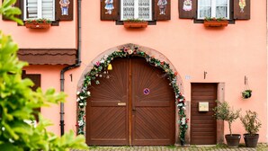 A building with a large wooden double door, a small window with a flower box, and a potted plant on the ground. - Gite "Au Lalli" – Kaysersberg Vignoble: Alsatian Charm in a Renovated 1721 House (Kaysersberg Vignoble)