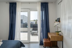 Ein Schlafzimmer mit Bett, Schreibtisch, Stuhl, einem Fenster mit Vorhängen und Blick auf ein Gebäude.