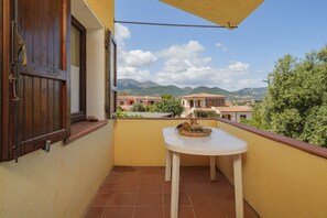 Ein Balkon mit Tisch, ein Fenster mit Holzjalousien und Blick auf entfernte Gebäude und Berge.