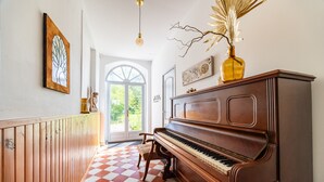 Un piano de cua en una habitació amb un terra de pissos quadriculats, un radiador de fusta i un gran finestral amb vista a vegetació.