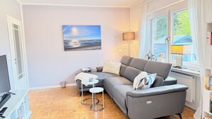 Uma sala de estar moderna com um sofá cinza, uma mesinha redonda, um quadro com uma paisagem praiana, uma luminária e uma estante de livros.