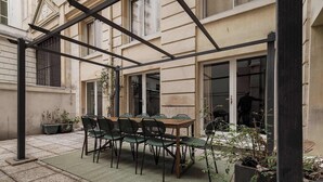 Un patio con una mesa y sillas de madera, plantas en macetas y una estructura de pérgola de metal.