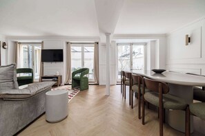 A modern living room with a sofa, a dining table with chairs, a TV mounted on the wall, and large windows. - Modern 10PAX/4BDR apartment in the center (Paris)