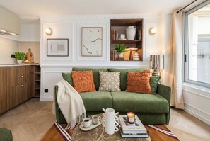 A green sofa with decorative pillows, a coffee table with a tray and cups, a bookshelf with books and decorative items, and a window with curtains. - 4PAX flat in the center of Paris (Paris)