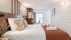 A hotel room with a wooden headboard, a large bed with white and floral bedding, a brown throw blanket, a bedside table with a vase and candle, and a view of the outdoors through a window. - 6PAX Spacious Apartment in the center (Paris)