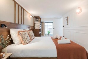 Ein Hotelzimmer mit einem hölzernen Kopfteil, einem großen Bett mit weißer und floraler Bettwäsche, einer braunen Tagesdecke, einem Nachttisch mit einer Vase und Kerze sowie einem Blick ins Freie durch ein Fenster.