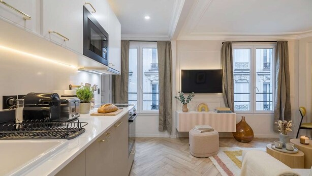 Una cocina moderna con horno integrado, microondas y cafetera. Hay una zona de comedor con mesa y sillas, y un amplio ventanal con cortinas.