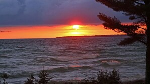 Una puesta de sol sobre el océano con olas, un árbol en primer plano y una costa distante.