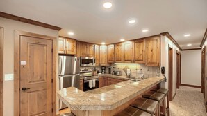 A kitchen with wooden cabinets, stainless steel appliances, a granite countertop, and a bar stool area. - Pool, Hot Tub, Complimentary Shuttle to Slopes, Great location! (Avon)