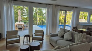 A spacious living area with a sofa, chairs, a small table, and a view of a swimming pool through large windows. - Hampton Bays Traditional with a heated pool, plenty of privacy, minutes to... (Hampton bays)