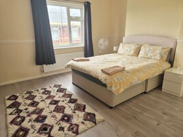 Un dormitorio con cama, mesita de noche, lámpara de pie, ventana con cortinas y una alfombra estampada.
