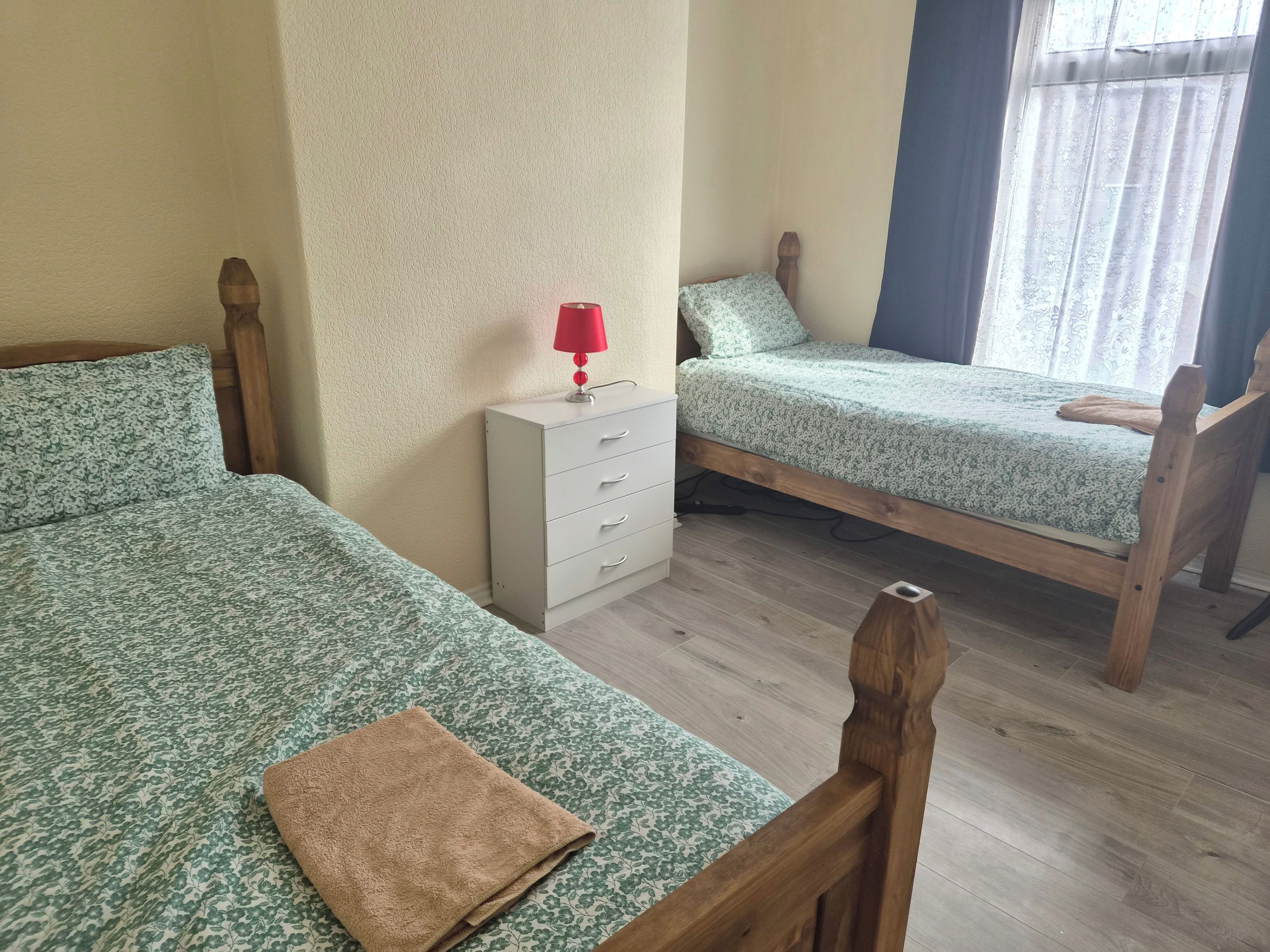 A room with two wooden beds, a white bedside table with drawers, a red lamp, and a window with curtains.