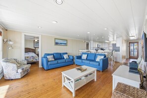 Uma sala de estar ampla com piso de madeira, dois sofás azuis, uma mesa de centro branca e uma cozinha ao fundo.
