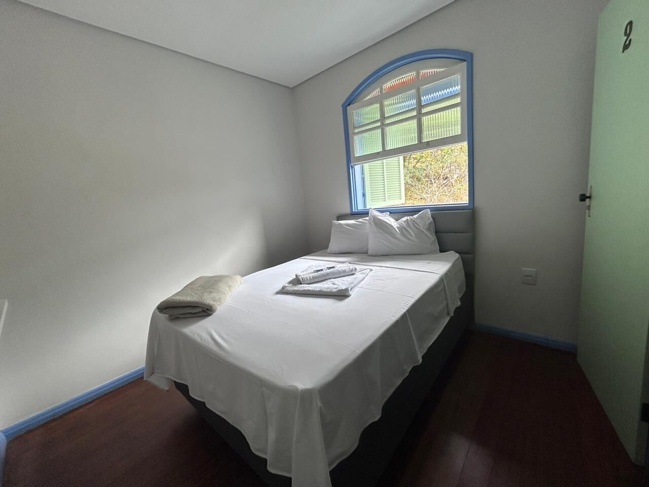 A single bed with white linens, a window with blue trim, and a door with a number.