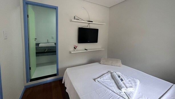 Habitación de hotel con cama, televisor montado en la pared, una pequeña estantería con un control remoto y un baño visible a través de una puerta abierta.