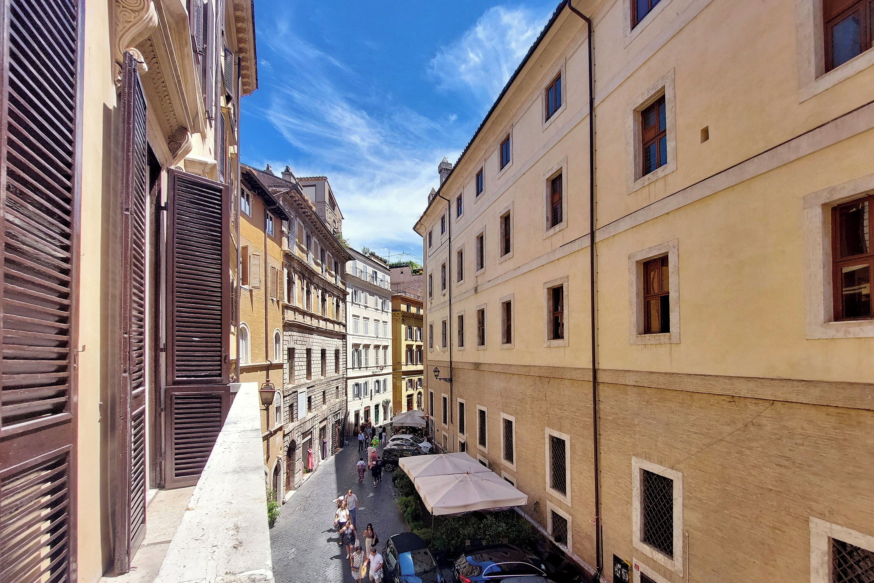 Una calle estrecha bordeada por edificios históricos con contraventanas, un café con terraza y coches aparcados.