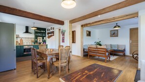 Amplia sala de estar con una mesa de comedor de madera, sillas y un sofá. Se ve una cocina al fondo con gabinetes verdes y una lámpara colgante.