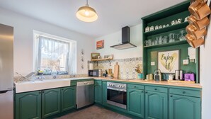 Una cocina con gabinetes verdes, lavavajillas, horno, microondas y una ventana con cortinas translúcidas.