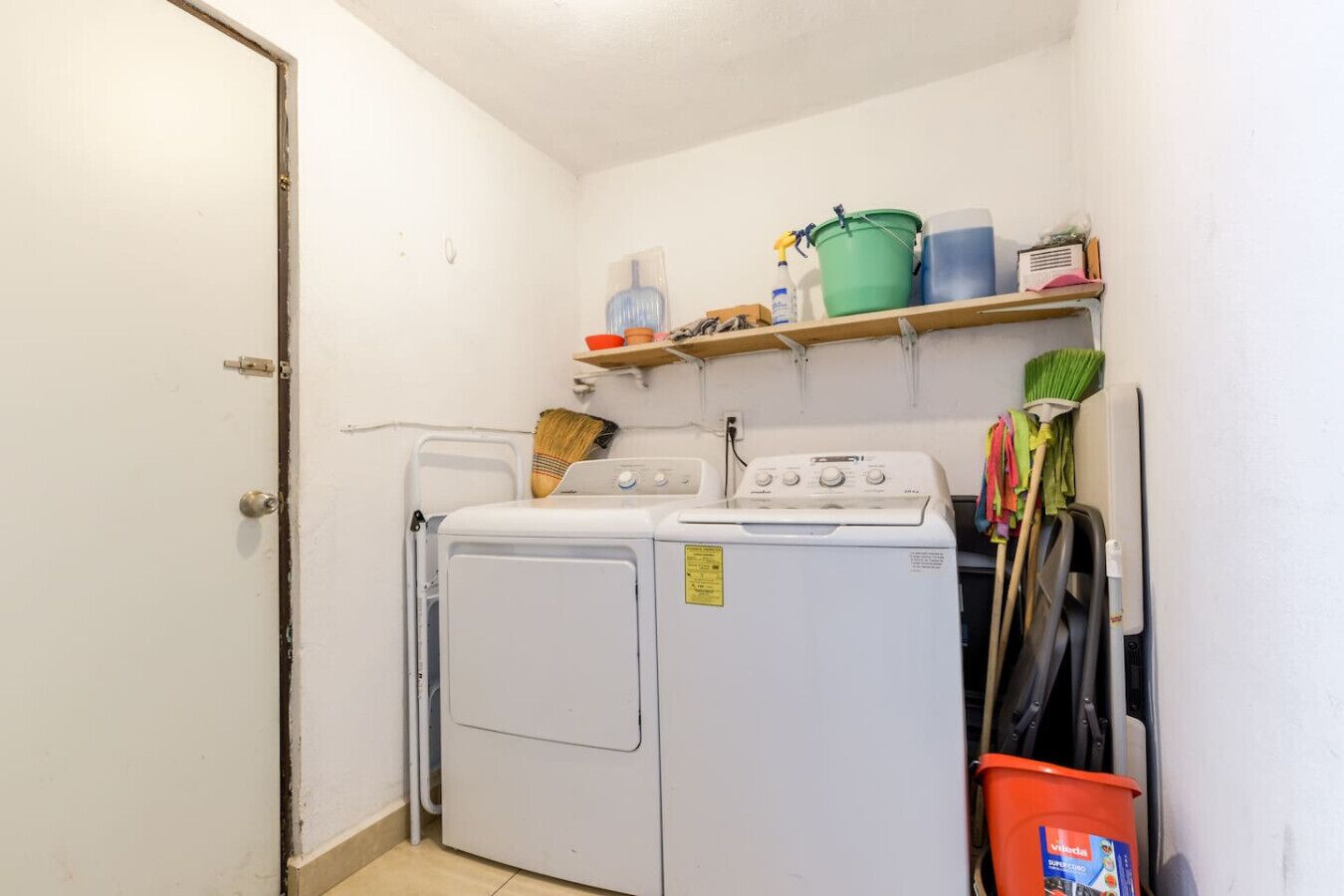 Una sala de bugaderia amb rentadora i assecadora, una escombra, un galleda i una prestatgeria amb productes de neteja.
