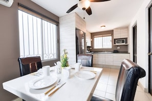 Un coin repas avec une table dressée pour deux personnes, un ventilateur de plafond, une cuisine en arrière-plan et une fenêtre avec des stores.