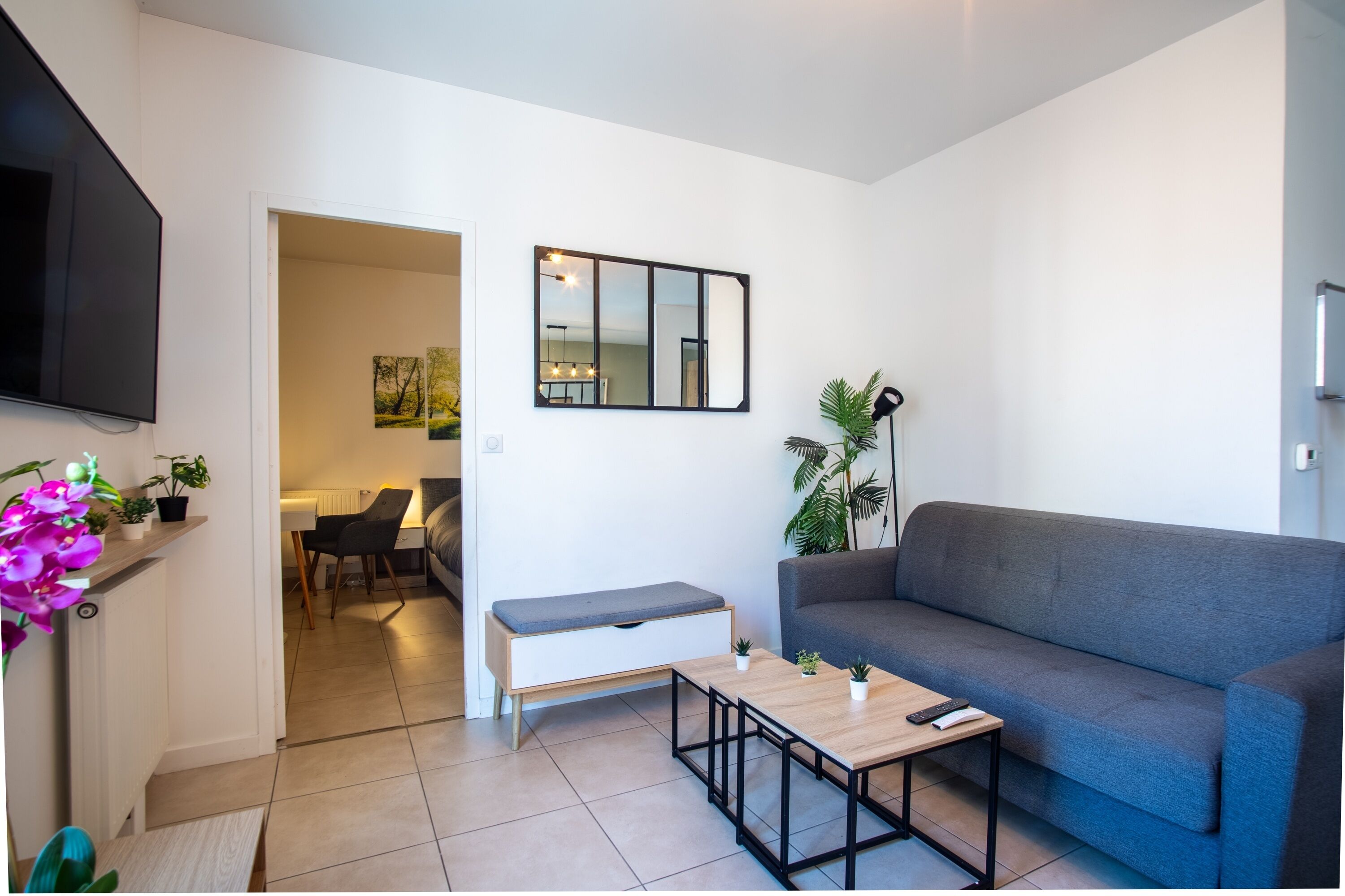 A modern living room with a grey sofa, a wooden coffee table, a bench, a TV, and a dining area with chairs and a table.