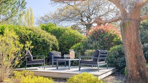 Een terras met tuinmeubilair, omringd door weelderige beplanting en een grote boom.