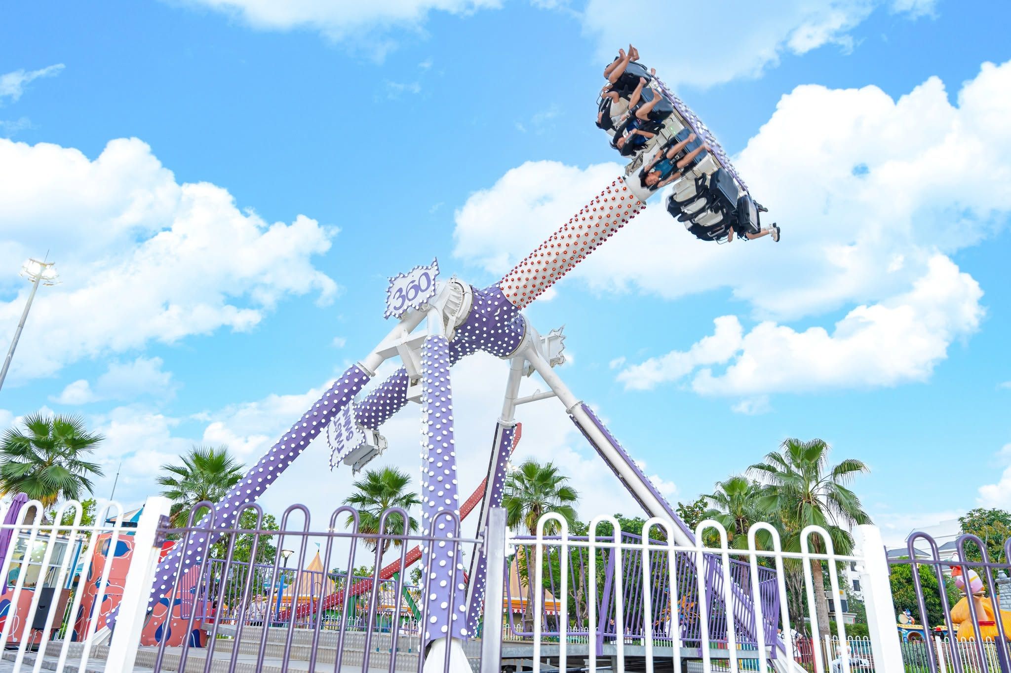 Una gran atracción de feria con una estructura mecánica en forma de brazo, rodeada por una valla blanca y palmeras.