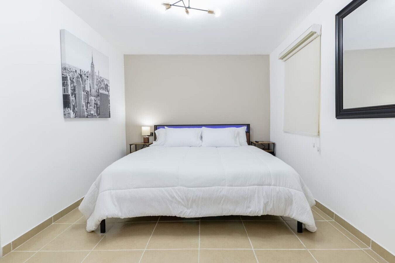 A hotel room with a large bed, a cityscape painting, a mirror, and a ceiling light fixture.