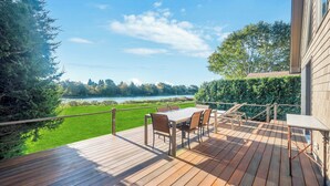 Um deck de madeira com mesa e cadeiras, com vista para um lago e rodeado por vegetação.