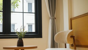 A room with a bed, a small round table with a vase of flowers, a window with curtains, and a chair with a woven headboard. - Moc Vien Homestay Train Street (Hanoi)