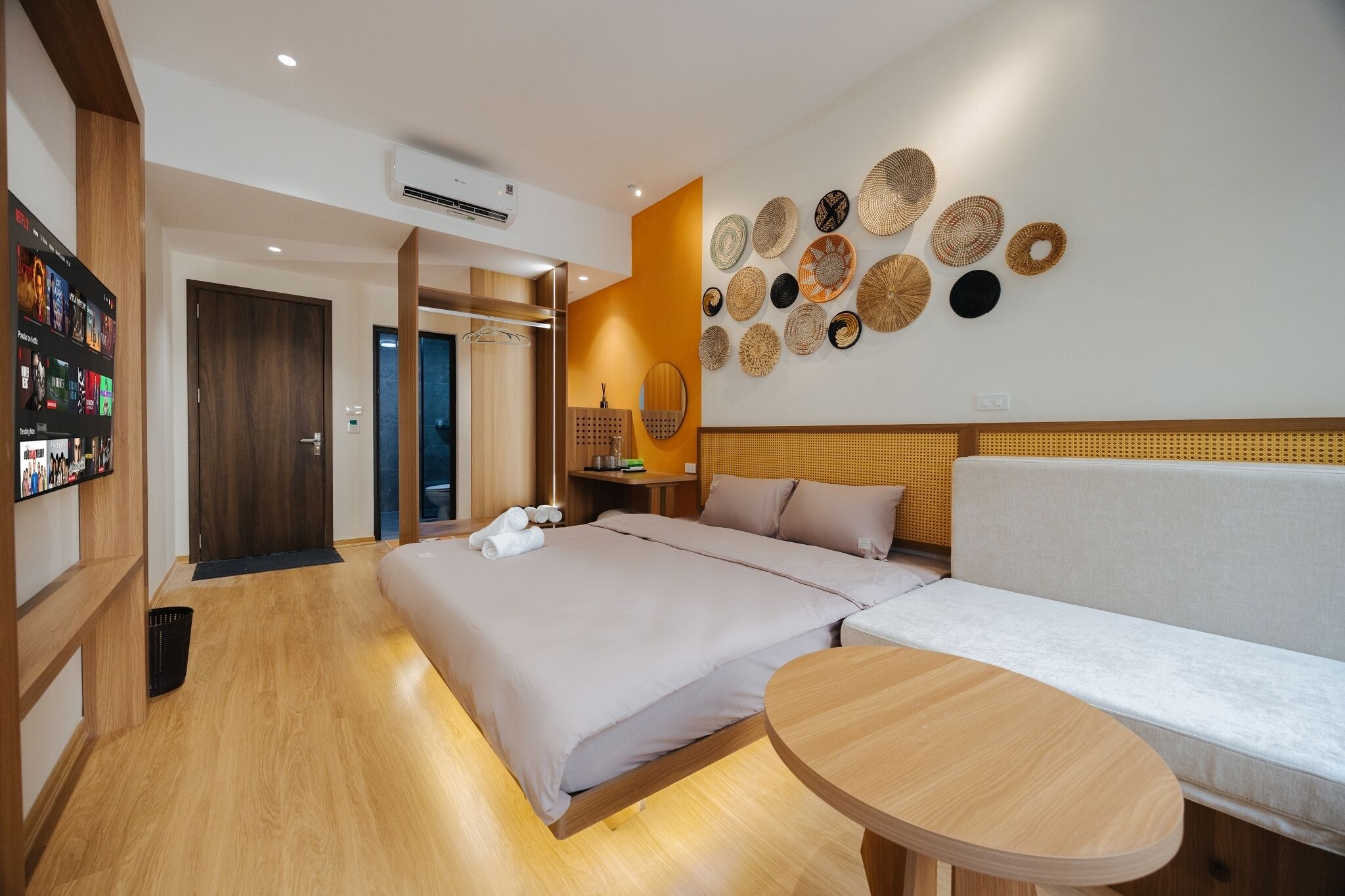 A hotel room with a bed, a sofa, a round table, a wall with woven baskets, and a hallway with a wooden floor and a door.