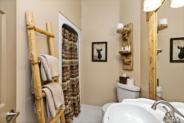 Un baño con un toallero de madera, cortina de ducha estampada, inodoro, lavamanos y estantes en la pared.