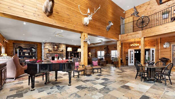A spacious room with a grand piano, a dining table with chairs, and a high ceiling with wooden beams and mounted animal heads. - Whitestone Ranch | Luxury Lakefront Estate Near Muskoka (Sleeps 24) (Dunchurch)
