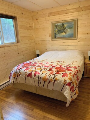 A wooden cabin bedroom with a bed, a window, a painting on the wall, and a nightstand.