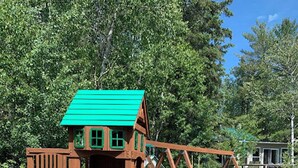 A wooden play structure with a slide and climbing area, set in a grassy area with trees in the background.