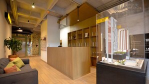 Un área de recepción con un mostrador de madera, una vitrina con una maqueta de un edificio y un sofá con cojines.