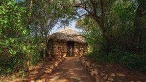 A stone hut with a thatched roof, surrounded by dense foliage and a dirt path leading to it. - Private Bush Camp With Pool Cottages Tents - 2332 (Bisil)