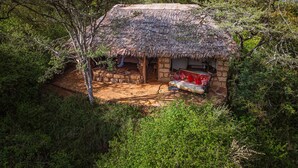 A thatched-roof hut with a stone wall, a sofa with cushions, and a small table. - Private Bush Camp With Pool Cottages Tents - 2332 (Bisil)