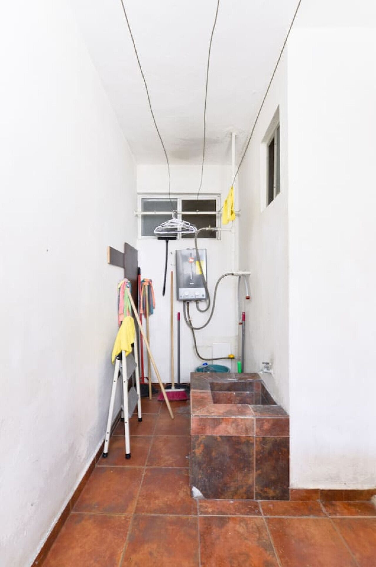 Um corredor estreito com piso de azulejos, uma janela e um espaço de serviço com materiais de limpeza.