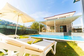 Une piscine avec des chaises longues et un parasol, un espace extérieur couvert et un bâtiment doté d’une façade en verre.