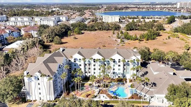 Un hotel de varios pisos con piscina, rodeado de vegetación y una extensa masa de agua al fondo.