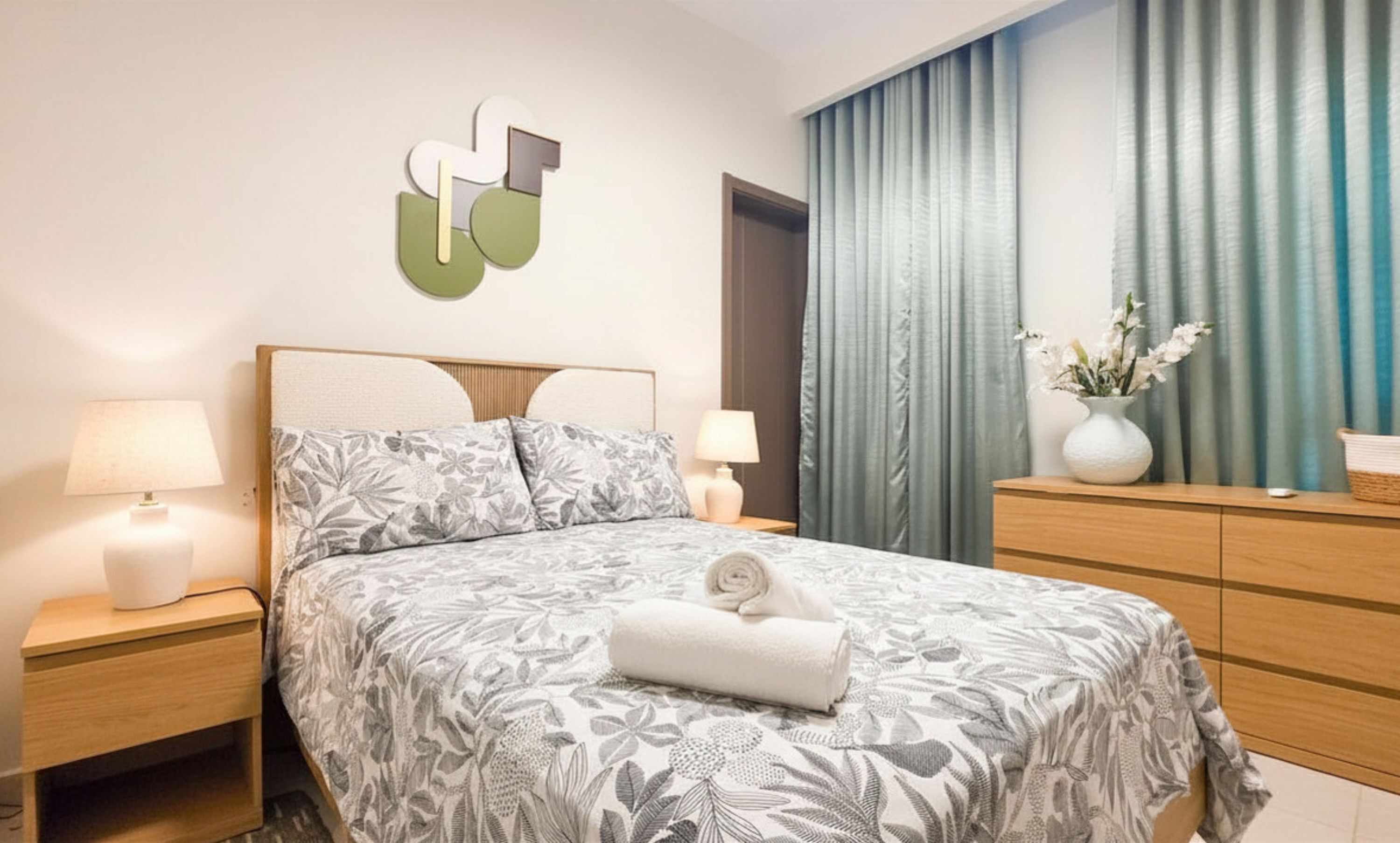 Una cama bien hecha con colcha estampada floral, dos lámparas de noche, un cuadro abstracto en la pared, un florero con flores y un aparador de madera.