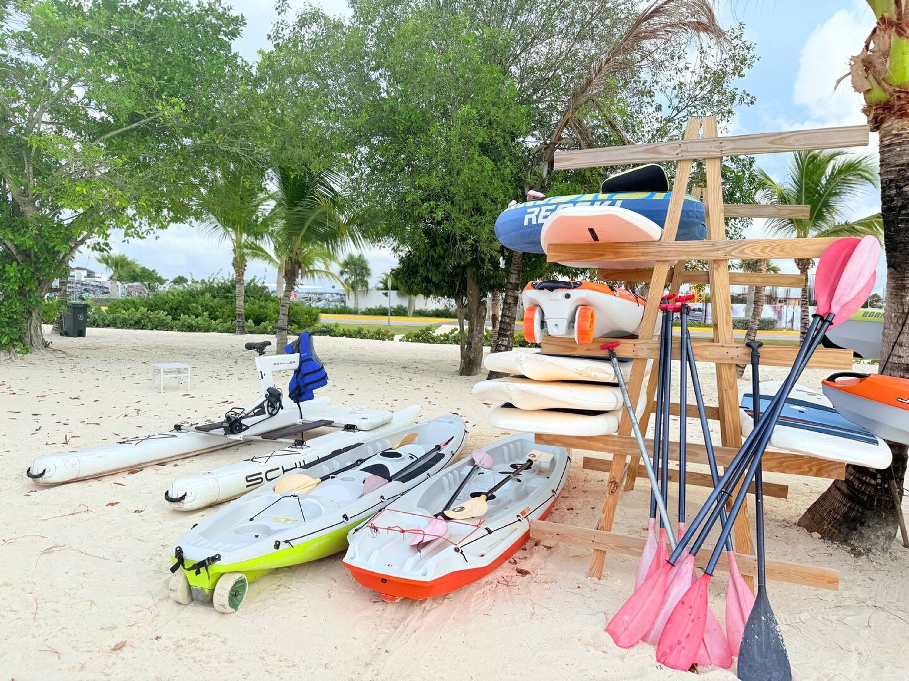 En strand med kajakker og paddleboards, et træstativ med årer og et lille bord med en stol.