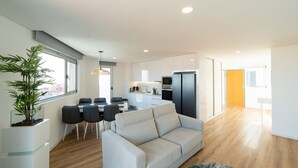 Ein modernes Wohnzimmer mit Sofa, Esstisch, Stühlen, einer Küchenzeile inklusive Geräte und einem großen Fenster.