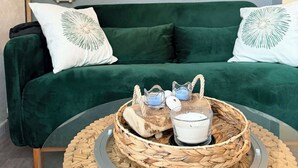 Ein grünes Sofa mit weißen Kissen, ein runder Glastisch mit einem gewebten Tablett und eine Wandleuchte.