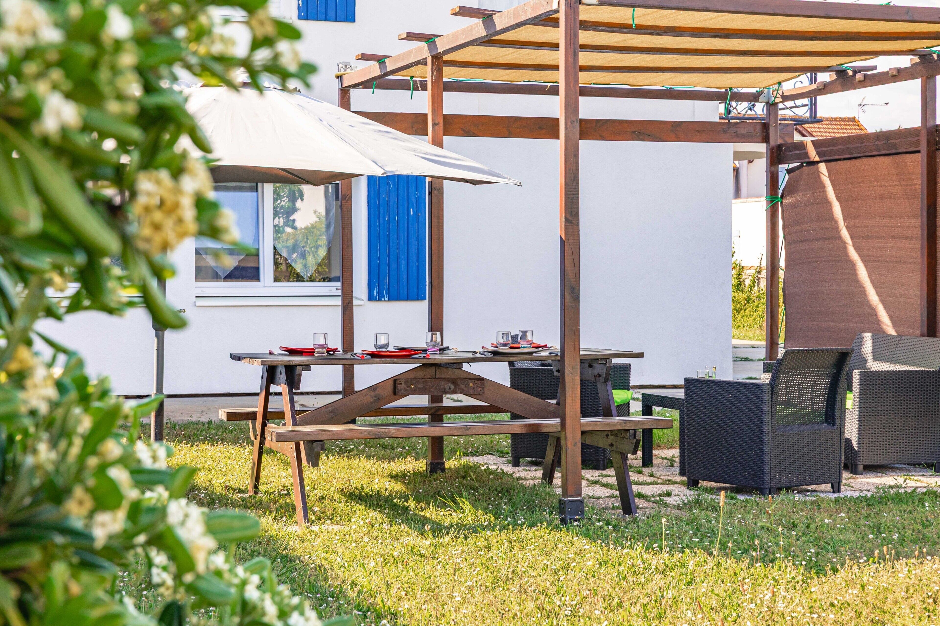 Eine Terrasse im Freien mit einem Holztisch, Stühlen, einer Pergola und einem weißen Gebäude im Hintergrund.