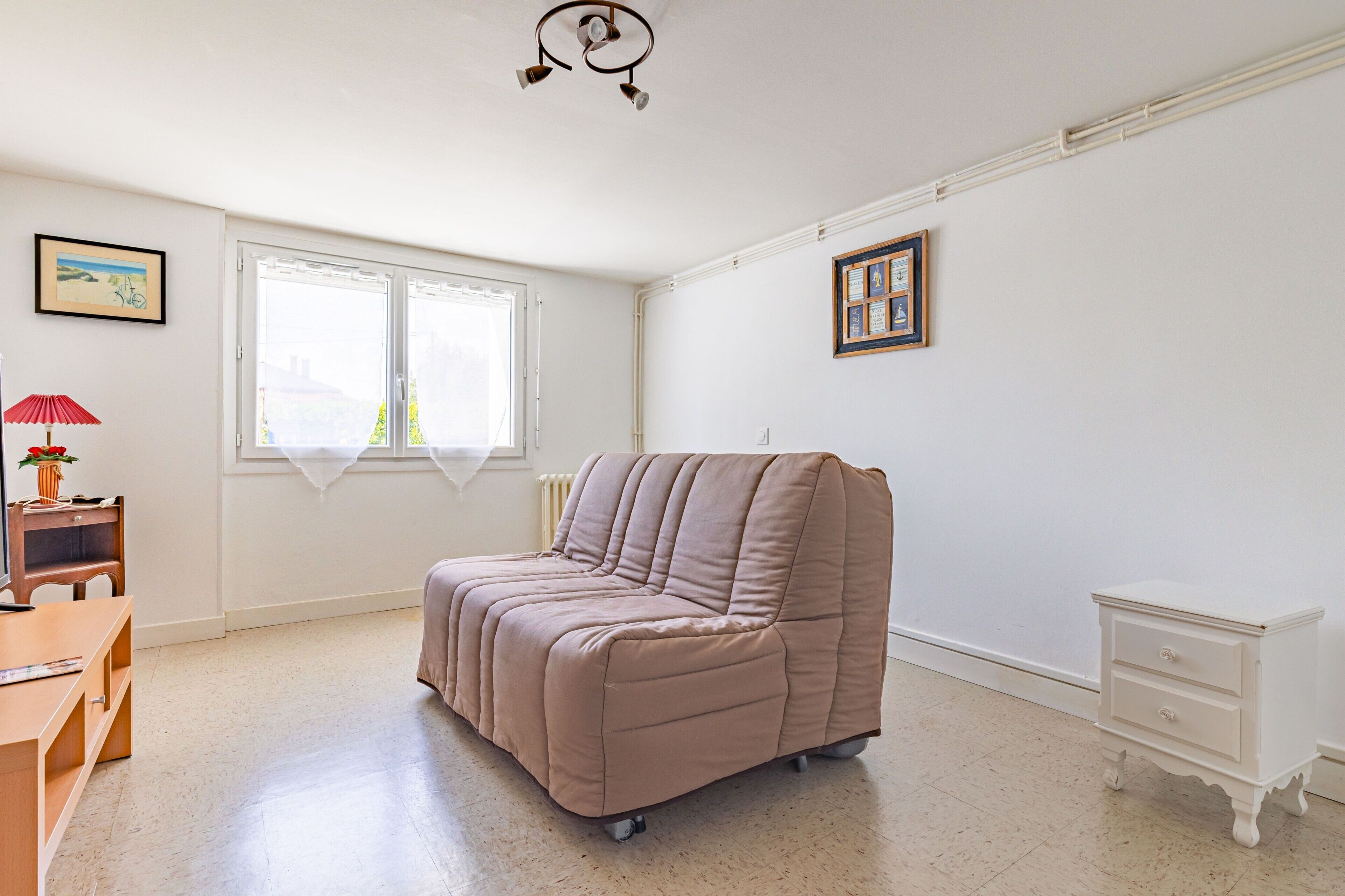 A spacious room with a beige sofa, a small wooden desk, a white nightstand, and a window with white curtains.