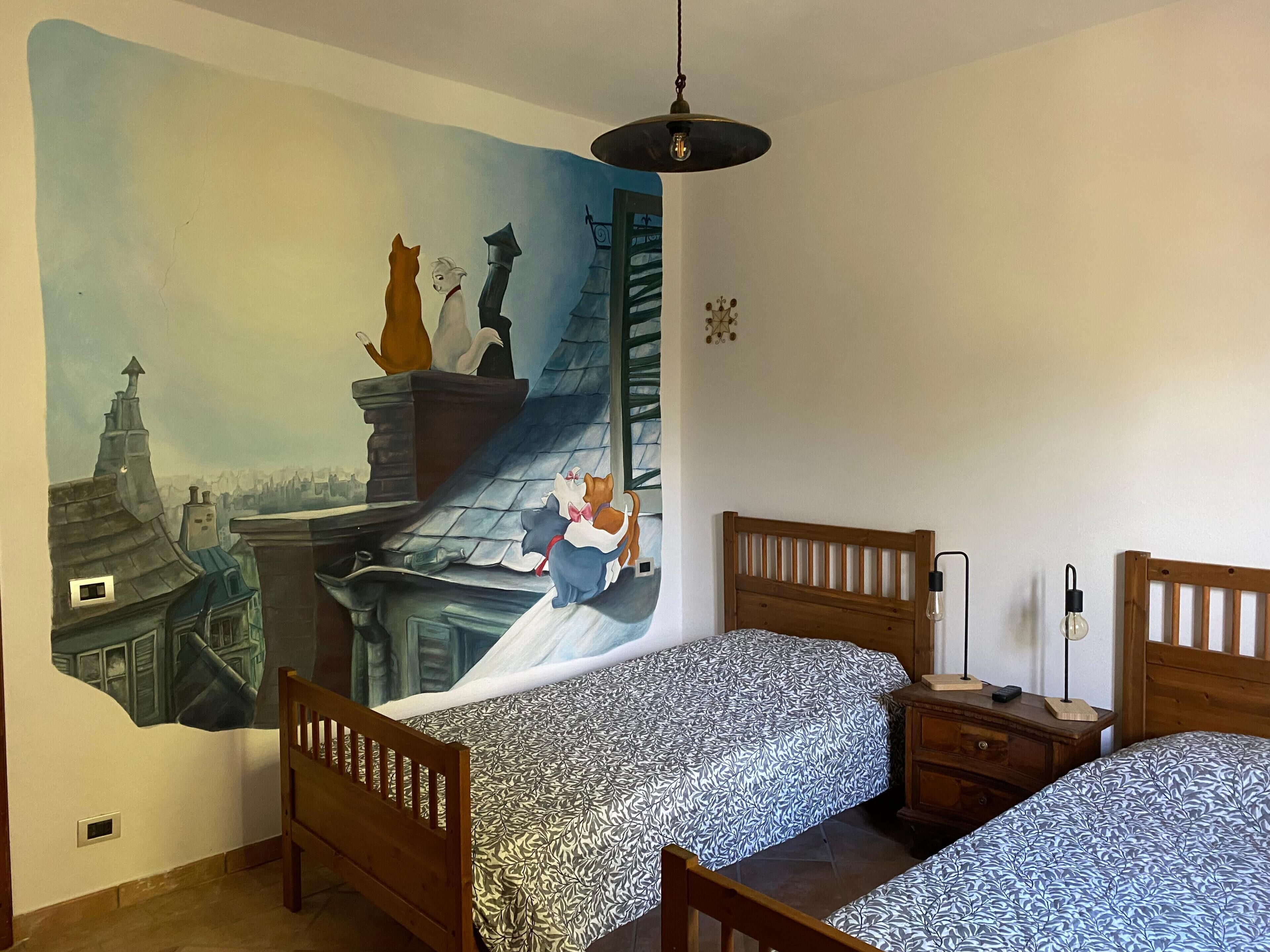 Um quarto com duas camas de madeira, um mural na parede, um lustre e uma mesa de cabeceira com abajur.